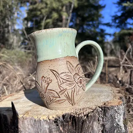 Hand thrown cup featuring raw speckled clay with hand carved daffodils and daisies. Finished with a green drippy glaze. 