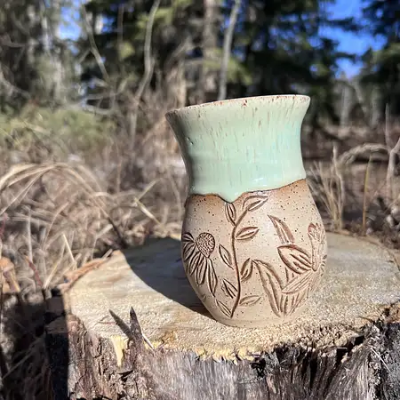 Hand thrown cup featuring raw speckled clay with hand carved daffodils and daisies. Finished with a green drippy glaze. 