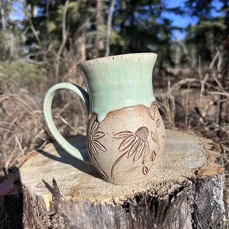 Hand thrown cup featuring raw speckled clay with hand carved daffodils and daisies. Finished with a green drippy glaze. 