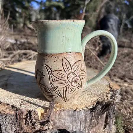 Hand thrown cup featuring raw speckled clay with hand carved daffodils and daisies. Finished with a green drippy glaze. 
